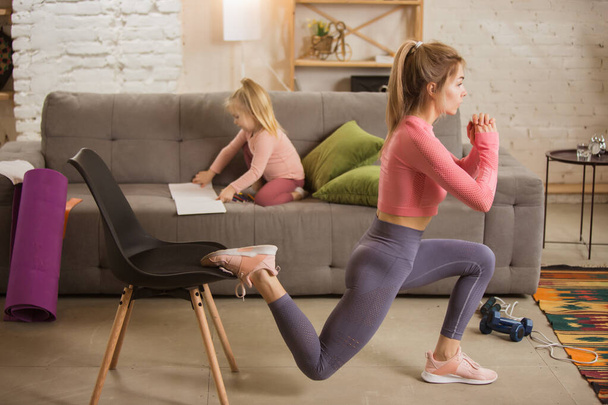 Young woman exercising fitness, aerobic, yoga at home, sporty lifestyle. Getting active with her child playing, home gym. - Foto, imagen