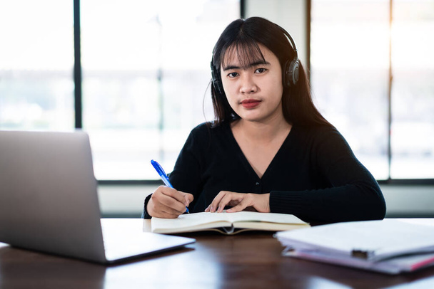 Lächelnde asiatische Studentin trägt drahtlose Kopfhörer schreiben auf dem Notizbuch, um Sprache online zu lernen und dem Dozenten zuzuhören, Webinar per Videoanruf E-Learning zu Hause, Fernstudium - Foto, Bild