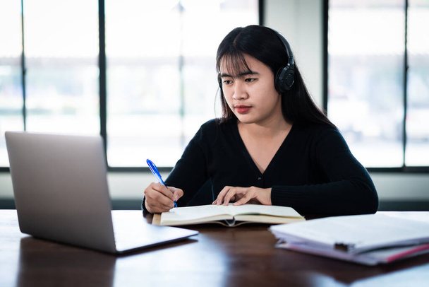 Smiling Asian girl student wears wireless headphones write on the notebook to study language online watch and listen to the lecturer, webinar via video call e-learning at home, distance education - Photo, Image