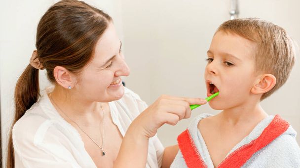Young smiling mother brushing and cleaning teeth of her little son with tothbrush. Concept of child dental hygiene and healthcare at home. Caring parents and kids at home. - Fotó, kép