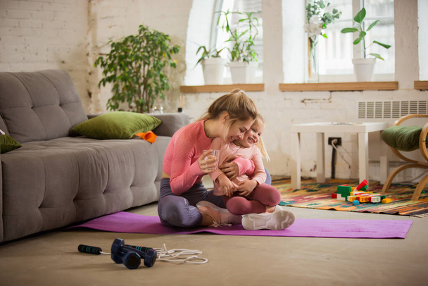 Young woman exercising fitness, aerobic, yoga at home, sporty lifestyle. Getting active while child, daughter playing with her, home gym. - Photo, Image
