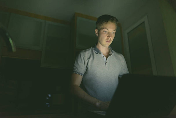 Πορτρέτο του νεαρού όμορφου άντρα που χρησιμοποιεί φορητό υπολογιστή στο σκοτεινό σαλόνι - Φωτογραφία, εικόνα