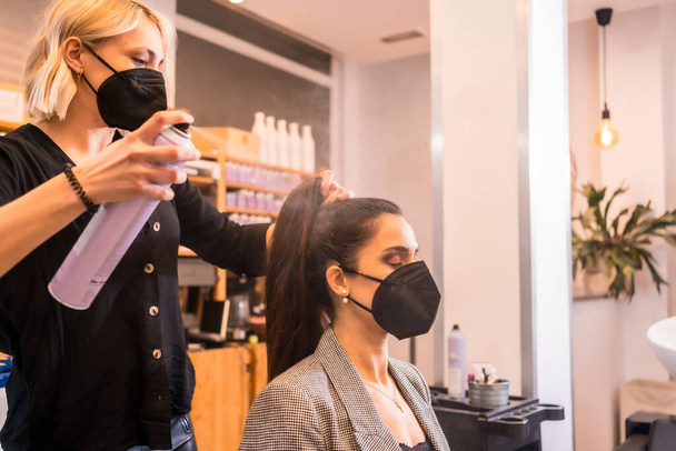 Hairdresser with face mask combing the client's hair with hairspray. Reopening with security measures of Hairdressers in the Covid-19 pandemic - Photo, Image