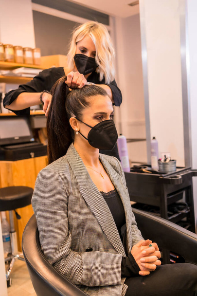 Caucasian hairdresser with mask finishing the hairstyle to the brunette client. Security measures of Hairdressers in the Covid-19 pandemic - Photo, Image
