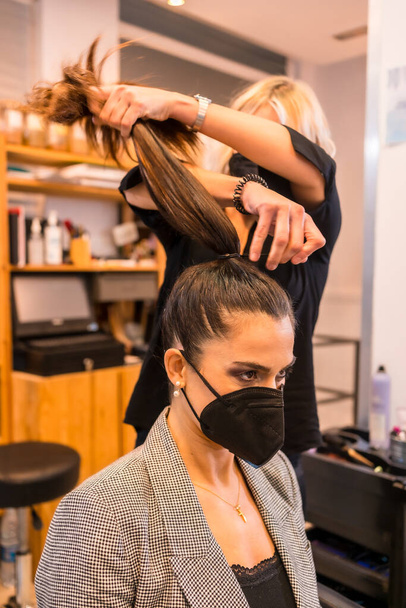Caucasian hairdresser with mask finishing the hairstyle to the brunette client. Security measures of Hairdressers in the Covid-19 pandemic - Foto, immagini