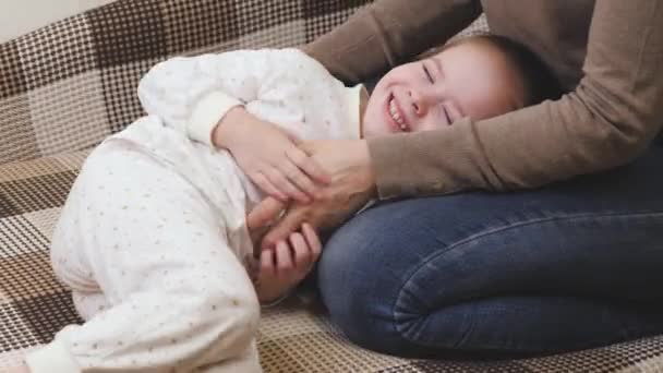 Το κορίτσι παίζει με τη μητέρα της στο παιδικό δωμάτιο στον καναπέ και γελάει. Ευτυχισμένη οικογενειακή ζωή. Ημέρα μητέρων. Το μωρό χαμογελάει ξαπλωμένο δίπλα στη μητέρα. Ο γονιός κάνει το μικρό παιδί να γελάει. - Πλάνα, βίντεο