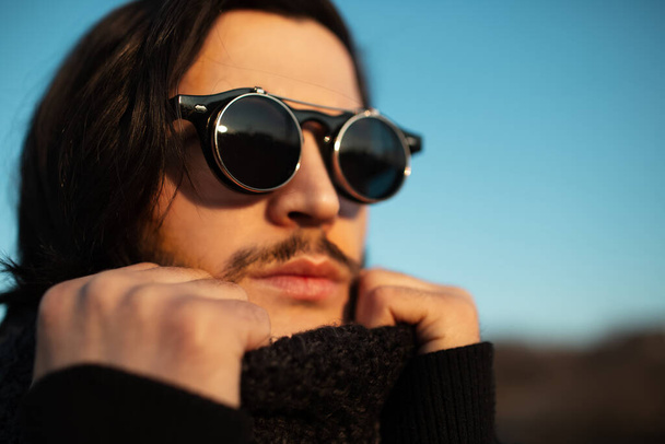 Egy közeli kép fiatal fickóról hosszú hajjal, aki hipszter kerek napszemüveget és sálat visel. Kék ég a háttérben. - Fotó, kép