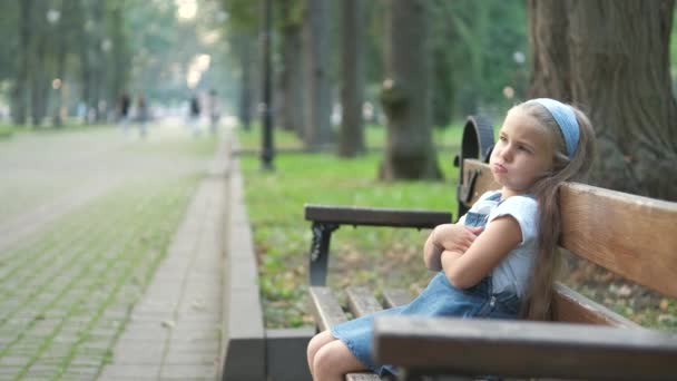 Petite enfant sérieuse assise seule sur un banc dans un parc d'été. - Séquence, vidéo