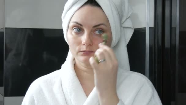 Mulher bonita com toalha na cabeça fazendo massagem com um rolo de jade para o rosto foreye área cabeça de pedra no banheiro. Ferramentas de beleza chinesas. Drenagem linfática massagem da pele contra o envelhecimento rugas - Filmagem, Vídeo