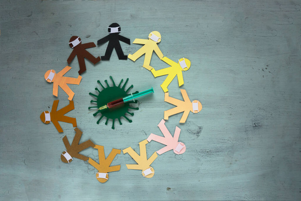 Différents mannequins de papier portant des masques et étant de différentes couleurs arrenged dans un cercle arround une seringue et un symbole de coronavirus - Photo, image