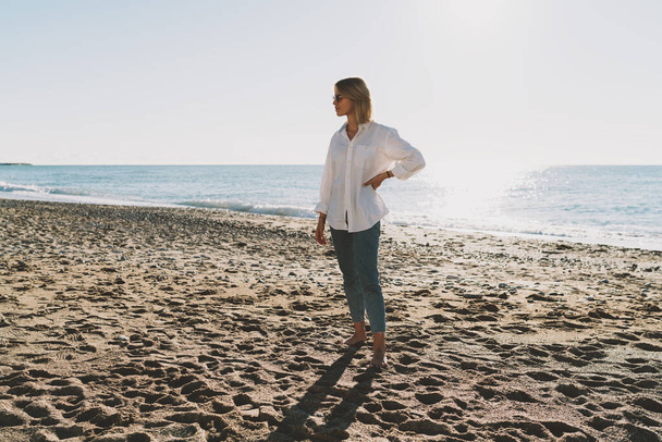 Full length portrait of an elegant blonde woman wearing white shirt and blue jeans looking aside while standing with the left hand on her waist at the beachside on a sunset time beside the sea water. - Photo, Image