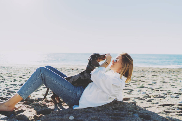 Happy dog owner female treating the pup while playing at the beachside. Blonde woman happy to have fun with her dog at the seaside on a sunny day. Hipster girl in casual outfit hugging the dog. - 写真・画像