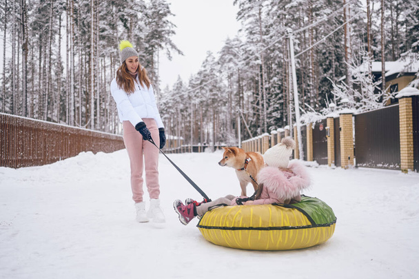 Madre feliz y niña linda en ropa interior de color rosa cálido caminar divertirse paseos inflable tubo de nieve con perro inu shiba rojo en el campo de invierno blanco nevado al aire libre. Deportes de familia actividades de vacaciones - Foto, imagen