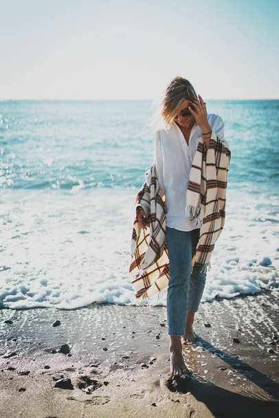 Retrato completo de una chica hipster rubia elegante con una manta sobre sus hombros caminando descalza el agua en una orilla del mar. Mujer joven en gafas de sol con estilo disfrutando de sus vacaciones en la playa. - Foto, Imagen