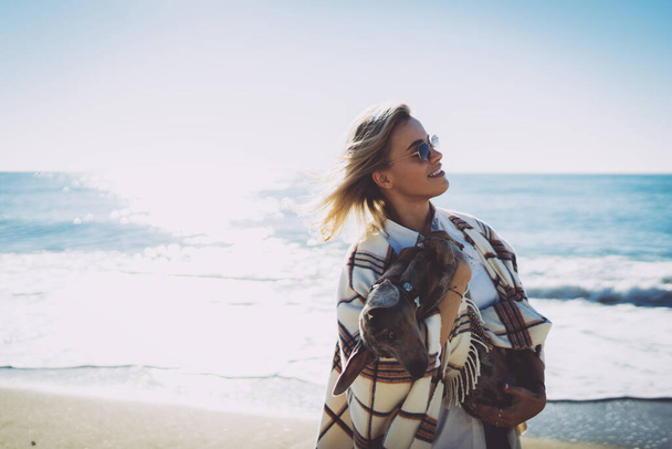Half length portrait of a charming blonde woman in sunglasses hugging lovely doggy while standing on a blurred beachside background. Attractive female enjoying walking the sea shore with her pet dog. - Photo, Image