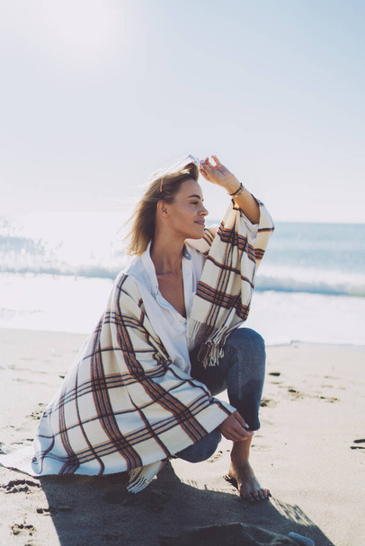Beautiful barefoot blonde woman wearing casual outfit taking off her sunglasses while sitting on haunches at seaside on a windy sunny day. Caucasian female with a blanket on her shoulders on a beach - Photo, Image