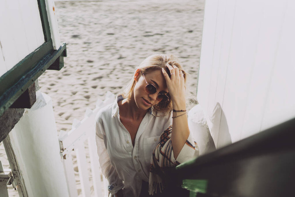 Attractive blonde Caucasian female in stylish sunglasses wearing white shirt with a blanket on her shoulders adjusting her hair with the hand while standing beside the wooden gate of a beach house.  - Foto, Bild
