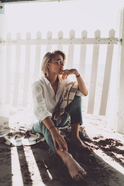 Ragazza bionda a piedi nudi premurosa seduta all'ombra accanto alla recinzione bianca sulla spiaggia. Bella donna caucasica in occhiali da sole con camicia bianca e jeans blu seduta sulla costa sabbiosa. - Foto, immagini