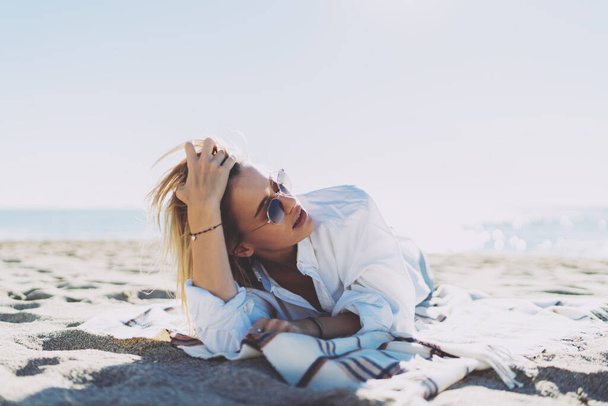 Портрет красивой белой женщины, которая прикасается к волосам и смотрит в сторону, наслаждаясь свободным временем на берегу моря. Блондинка-хипстер в современных солнцезащитных очках и модном наряде, расслабляющая на берегу моря - Фото, изображение