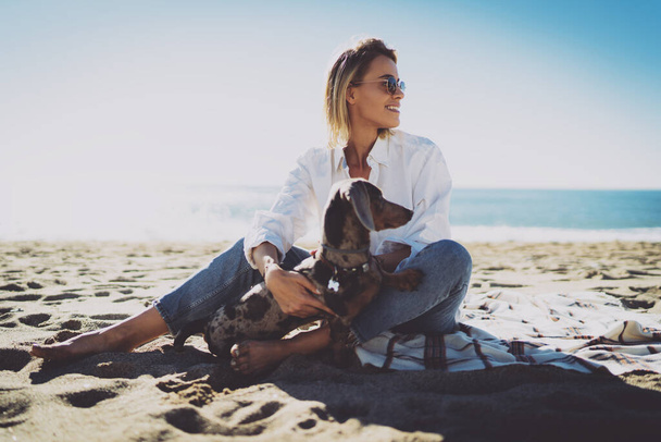 Beautiful blonde female in trendy sunglasses smiling aside while holding and cuddling her lovely pet dog. Cheerful young woman sitting on a blanket at the sandy beach side with her cute puppy. - Fotografie, Obrázek