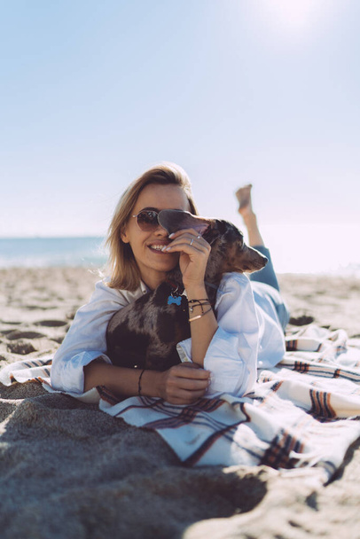 Cheerful female in casual outfit laughing and hugging dog while lying on a sand beach at the day off. Young woman smiling at the camera while relaxing with her pet dog on a warm sunny day outdoors. - Photo, Image