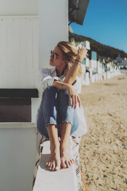Pensive blonde woman looking aside touching her hair while sitting on a parapet at the beachside. Model look female in sunglasses wearing white shirt and blue jeans spending sunny day on a beach. - Foto, Bild