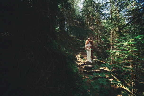 Gelukkig stijlvol stel pasgetrouwden in het groene bos op zomerdag. Bruid in lange witte jurk en bruidegom in rood pak knuffelen. trouwdag - Foto, afbeelding