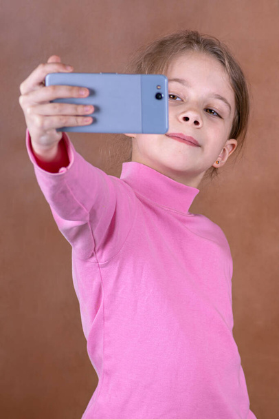 Portrait of a cheerful little girl taking a selfie on blurred brown background - Foto, Imagen