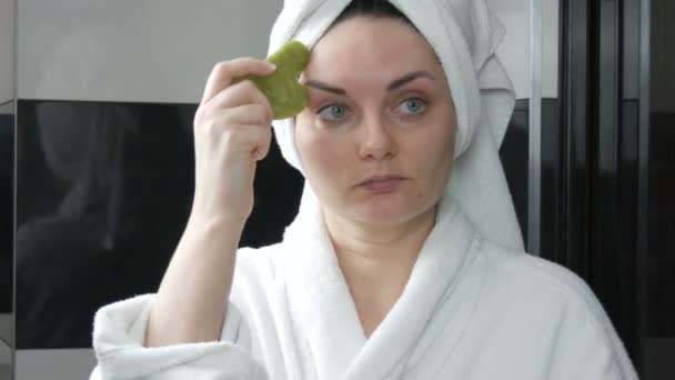 Kaunis nainen pyyhe päässä tekee hierontaa Jade kivi Kaavin Gouache otsa kasvot Hieronta kylpyhuoneessa. Kiinalaisia kauneustyökaluja. Imusolmukkeiden salaojitus ihon hieronta vastaan ikääntyminen ryppyjä - Materiaali, video