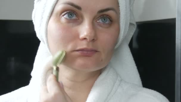 Гарна жінка з рушником на голові масаж з нефритовим роликом на обличчі природного зеленого каменю у ванній кімнаті. Китайські інструменти краси. Лімфатичний масаж шкіри від старіння зморшок. - Кадри, відео