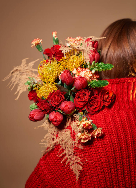 Ελκυστικό κορίτσι σε κόκκινο πουλόβερ κρατώντας χρωματιστό μπουκέτο από όμορφα τριαντάφυλλα, τουλίπες, γαρύφαλλα λουλούδια πίσω από την πλάτη της. Ημέρα του Αγίου Βαλεντίνου, Παγκόσμια Ημέρα της Γυναίκας ή έννοια της Μητέρας - Φωτογραφία, εικόνα