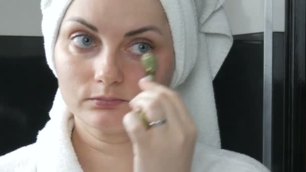 Krásná žena s ručníkem na hlavě při masáži s nefritovým válečkem na obličej přírodního zeleného kamene v koupelně. Čínské kosmetické nástroje. Lymfatická drenáž kůže masáž proti stárnutí vrásek - Záběry, video