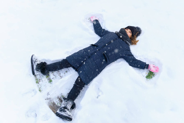 Κορίτσι παιδί βρίσκεται στο χιόνι και κάνει φτερά αγγέλου, λαμπερό φως του ήλιου και σκιές στο χιόνι, όμορφη φύση. - Φωτογραφία, εικόνα