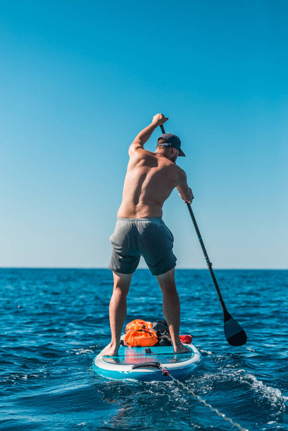 Young athletic man paddling on a SUP stand up paddle board in blue water sea in Montenegro - Photo, image