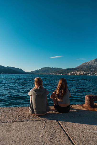 Achteraanzicht: Twee jonge vrouwen zitten aan de pier met uitzicht op de zee in Montenegro - Foto, afbeelding
