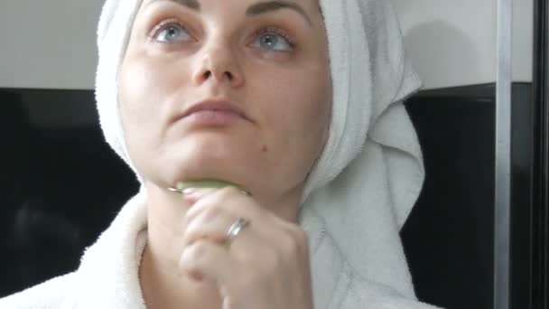 Piękna kobieta z ręcznikiem na głowie robi masaż jadeitowym wałkiem na twarz naturalnego zielonego kamienia w łazience. Chińskie narzędzia piękności. Masaż limfatyczny skóry przed starzeniem się zmarszczek - Materiał filmowy, wideo