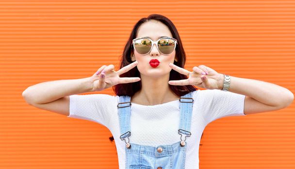 Beautiful brunette woman in sunglasses blowing lips kiss over orange background.  Outdoors. Young  woman. Street fashion. Red lipstick. - Photo, image