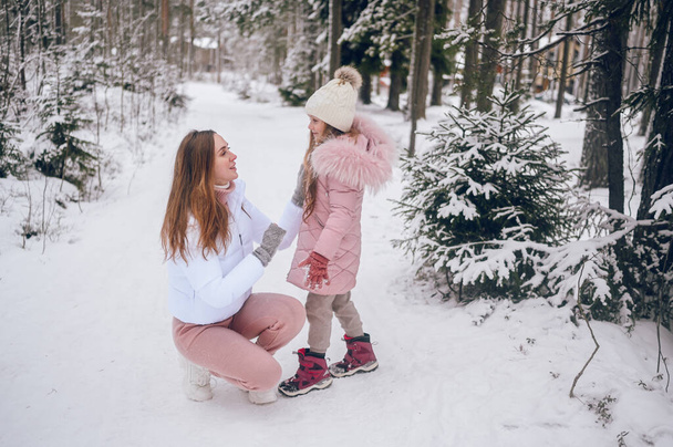 Ευτυχισμένη μητέρα και μικρό χαριτωμένο κορίτσι σε ροζ ζεστό outwear περπάτημα διασκεδάζοντας και αγκαλιάζοντας σε χιονισμένο λευκό κρύο χειμώνα κωνοφόρων δάσος με έλατο δάσος σε εξωτερικούς χώρους. Δραστηριότητες οικογενειακών αθλητικών διακοπών - Φωτογραφία, εικόνα