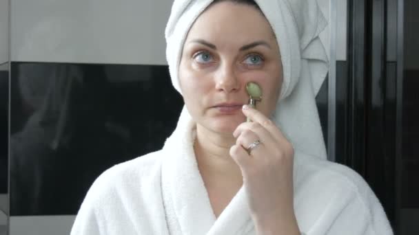 Hermosa mujer con toalla en la cabeza haciendo masaje con un rodillo de jade para la cara nasolabial pliegues de piedra en el baño. Herramientas de belleza chinas. Drenaje linfático masaje de la piel contra las arrugas del envejecimiento - Imágenes, Vídeo