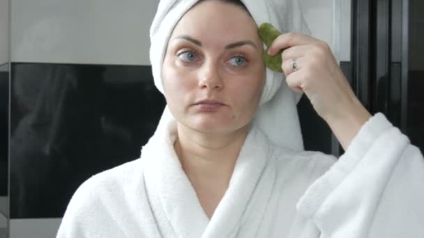 Bella donna con asciugamano sulla testa facendo massaggio con un raschietto di pietra di giada per guazzo fronte Massaggio in bagno. Strumenti di bellezza cinesi. Massaggio linfatico drenante cutaneo contro l'invecchiamento delle rughe - Filmati, video