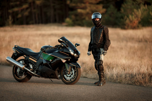 Conducteur de moto dans un casque et veste en cuir se tient sur la route à côté d'une moto de sport - Photo, image