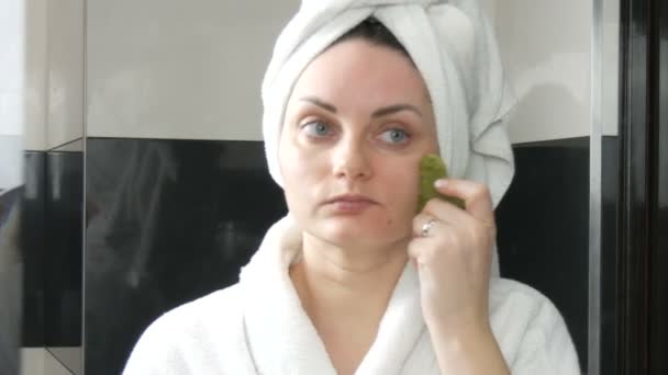 Kaunis nainen, jolla on pyyhe päässä tekemässä hierontaa jade-rullalla luonnollisen vihreän kiven kasvoille kylpyhuoneessa. Kiinalaisia kauneustyökaluja. Imusolmukkeiden salaojitus ihon hieronta vastaan ikääntyminen ryppyjä - Materiaali, video