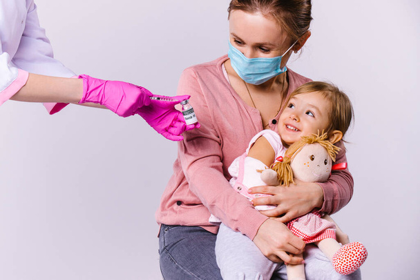 Mom and little girl are happy in a childrens clinic receive a vaccine against covid 19. White background and approach to young children. - 写真・画像