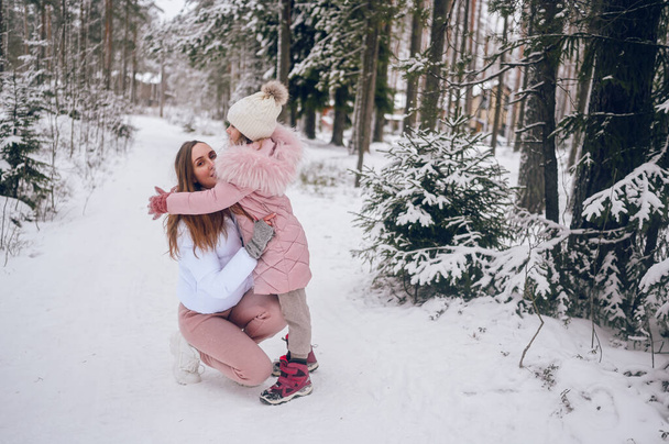 Mãe feliz e pequena menina bonito em roupas quentes rosa andando se divertindo e abraçando na floresta de coníferas de inverno frio branco nevado com madeiras de abeto ao ar livre. Desporto familiar atividades de férias - Foto, Imagem