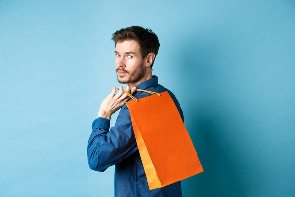 Rear view of happy man holding shopping back on shoulder and looking behind, standing on blue background - 写真・画像
