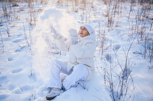 Щаслива літня старша зріла жінка в білому теплому одязі грає зі снігом на сонячній зимовій вулиці. Відпустка здорових людей відпочинок зимові заходи, концепція активного способу життя
 - Фото, зображення