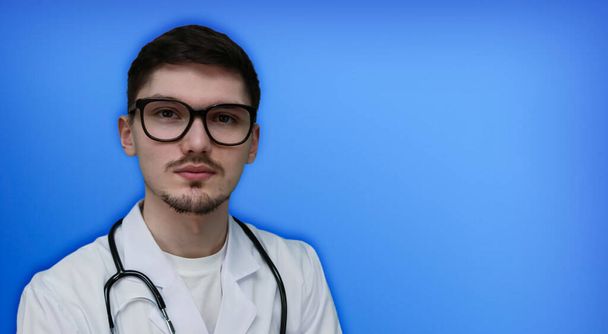 Портрет молодого лікаря в окулярах і стетоскоп зліва на синьому фоні з пробілом для тексту справа, крупним планом
. - Фото, зображення