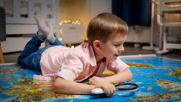 Ein kleiner lächelnder Junge liegt in seinem Zimmer auf dem Boden und erkundet die große Weltkarte durch eine Lupe. Konzept für Reisen, Tourismus und Kindererziehung. Kinder erkunden und entdecken - Foto, Bild