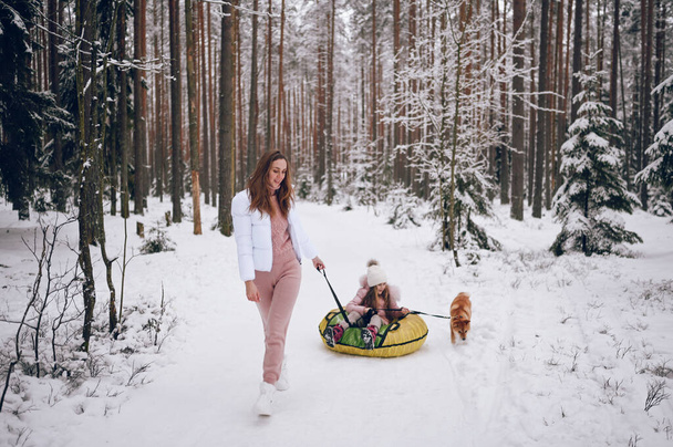 Madre feliz y niña linda en ropa interior de color rosa cálido caminar divertirse paseos inflable tubo de nieve con perro inu shiba rojo en nieve blanca fría bosque de invierno al aire libre. Deportes de familia actividades de vacaciones - Foto, imagen