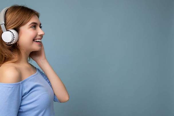 Бічно-профільна фотографія чарівної позитивної усміхненої молодої блондинки в синьому верхньому куті ізольованої на синьому фоні стіни в білих бездротових навушниках Bluetooth, що слухають прохолодну музику і
 - Фото, зображення